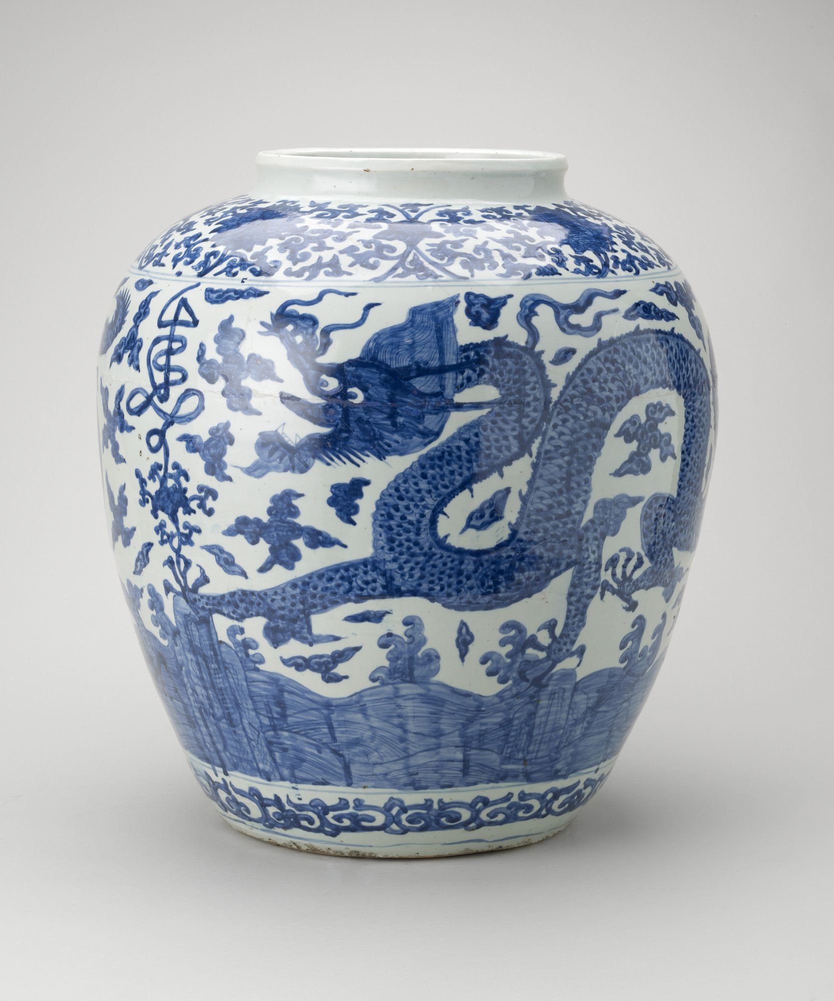 An ovoid-shaped Chinese Ming period porcelain jar painted in rich blue around the sides with two five-clawed dragons among clouds and with rocks and waves below. Round the shoulder a stylised shou (long life character) seems to grow out of the lotus scrol