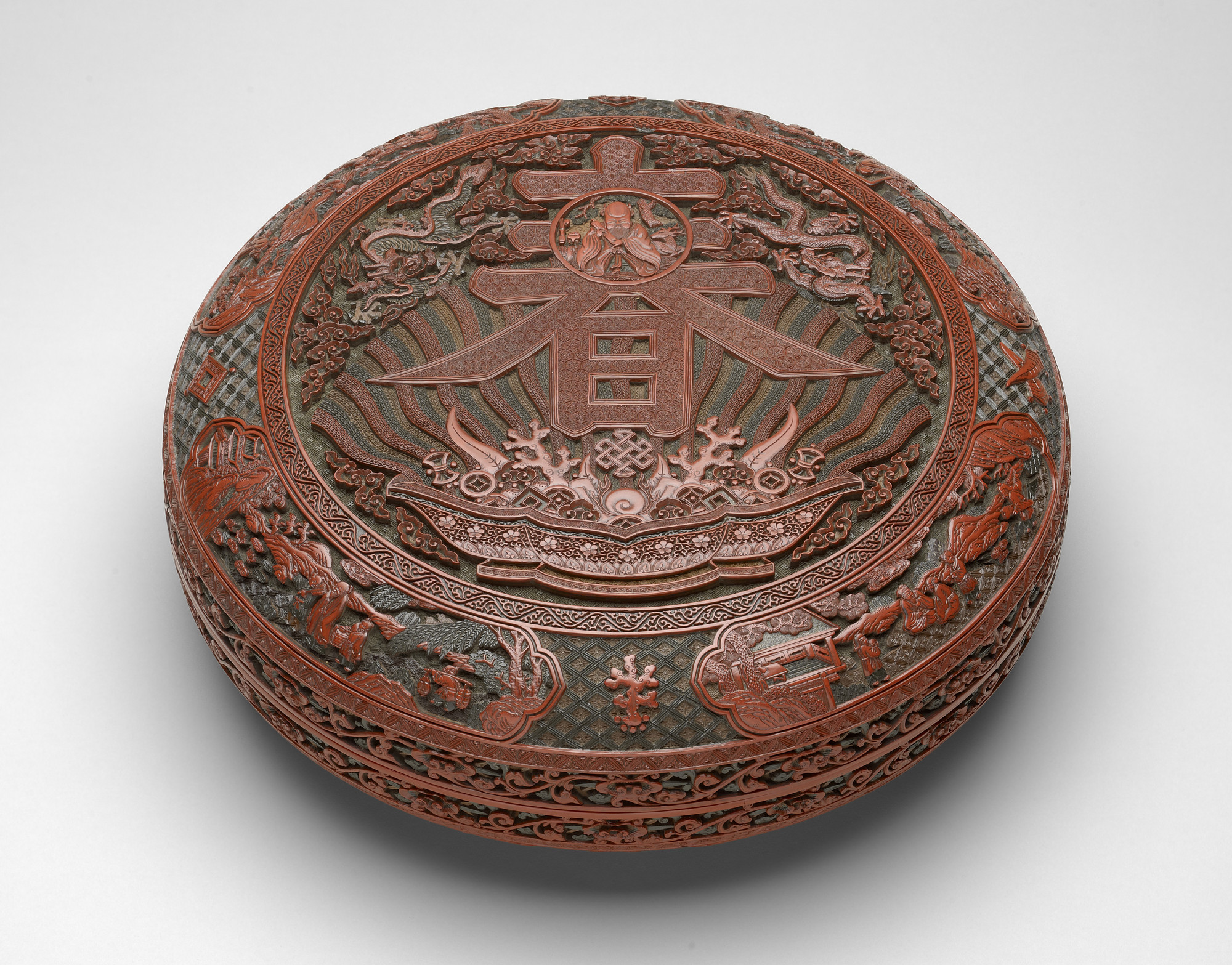 Large Imperial shallow box, with rounded sides and flanged rim, the matching cover with almost flat top. With many layers of ochre-yellow, green and red lacquer applied over a wood base. Carved on the top, a circular panel within a &lsquo;meander scroll&r