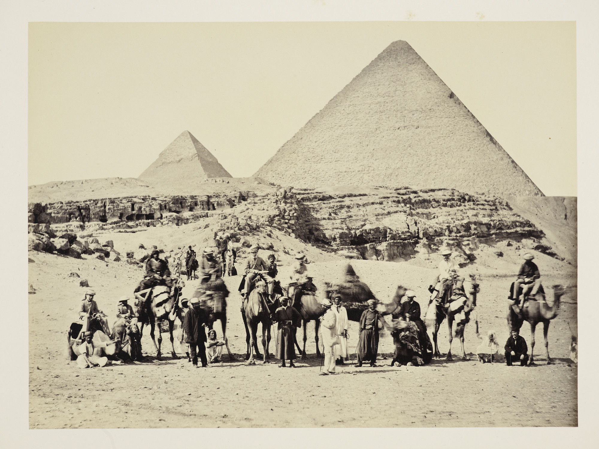 The Prince of Wales and his entourage on camels posing for camera in front of Pyramid of Cheops and Pyramid of Cephrenes, Giza, Cairo. The Prince is seated on the camel fifth from the left. The man in the white suit with a cigar, gazing up at the Prince, 