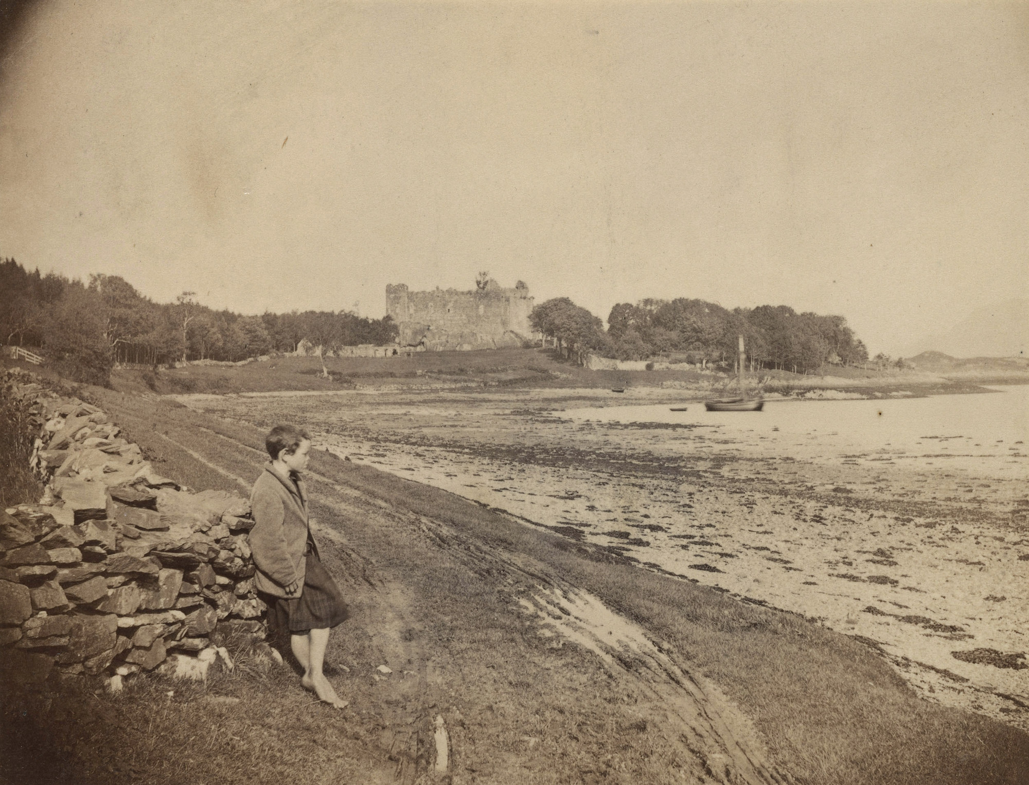 Photograph of a coastline with a young boy standing in right side profile as he leans against a&nbsp;low stone wall,&nbsp;looking out to&nbsp;sea. The boy wears three-quarter length trousers and is barefoot. In the background stands Dunstaffnage Castle on