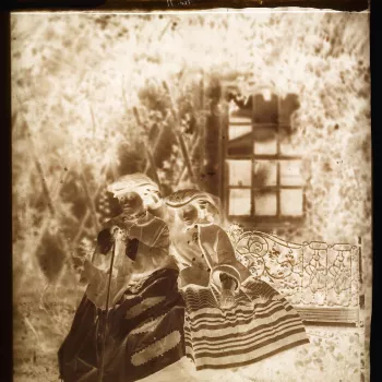 Glass plate negative of a full length portrait of Victoria, Princess&nbsp;Royal&nbsp;and Princess Alice. The sisters&nbsp;are seated together on a decorative ironwork garden seat. The Princess Royal holds a walking stick with Princess Alice leaning on her