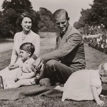 Photograph of HM The Queen (b. 1926) seated on the grass within the gardens of Clarence House. In front of her is seated HRH Prince Charles (b. 1948). HRH The Duke of Edinburgh (b. 1921) is seated on the Queen's left and looks at HRH Princess Anne (b. 195