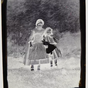 Glass plate negative of a full length double portrait of the sisters, Victoria-Alice and Mary Symons. The young girls stand in front of a wall and face the camera front on. The younger girl holds a doll. They both wear patterned dresses, light colour stoc